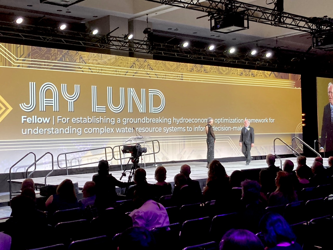 AGU 2022 Fellow, Jay Lund, walks across the stage with a description of his outstanding achievement projected on the screen in the background. Text says, "JAY LUND. Fellow | for establishing a groundbreaking hydroeconomic optimization framework for understanding complex water resource systems to inform decision-making." PC Alvar Escriva-Bou (Twitter: @AlvarEscriva); shared with permission.