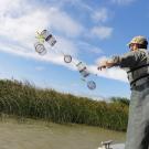 PC contributed by John Durand. Image: Person throwing a trap in the air toward the water; the image is taken such that the horizon is slanted and the in-action activity takes up most of the image. 